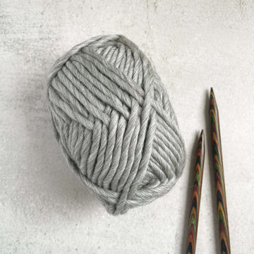 A ball of super chunky merino yarn in a natural grey colour with the pointy ends of two knitting needles.